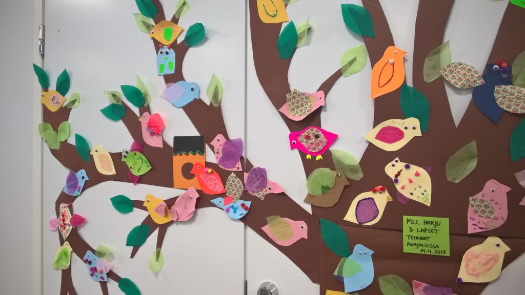 Kuvassa seinään kiinnitetty puu, jonka oksilla on värikkäitä paperisia lintuja